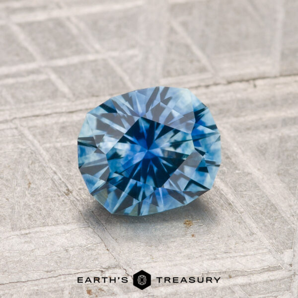 1.33-Carat Montana Sapphire (Heated) A 1.33-carat heated Montana sapphire with a geometric cut rests on a textured gray surface. The "Earth's Treasury" logo appears at the bottom of the image.