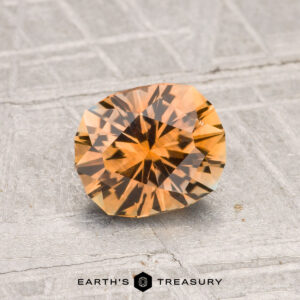 A 1.24-carat heated Montana sapphire with a faceted orange hue rests on a textured gray surface, with "EARTH'S TREASURY" visible at the bottom.