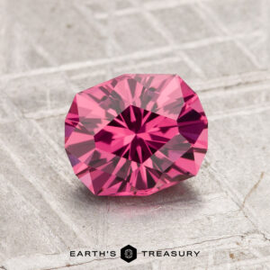 A 1.52-carat Tanga Garnet gemstone, cut with facets and pink in color, rests on a textured gray surface with the "Earth's Treasury" logo and text displayed at the bottom.