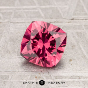 A vibrant 1.51-carat Tanga Garnet with a square cushion cut rests on a textured gray surface, with the "Earth's Treasury" logo visible at the bottom.