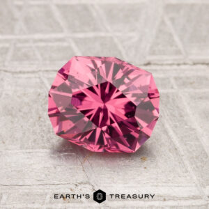 A 1.47-carat Tanga Garnet, round and faceted with a pink hue, is displayed on a textured gray surface with "Earth's Treasury" at the bottom.