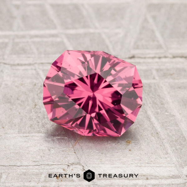 1.47-Carat Tanga Garnet A 1.47-carat Tanga Garnet, round and faceted with a pink hue, is displayed on a textured gray surface with "Earth's Treasury" at the bottom.