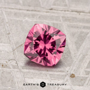 A 0.97-carat Tanga Garnet, cushion-cut and pink, is displayed on a textured gray surface with "Earth's Treasury" at the bottom of the image.