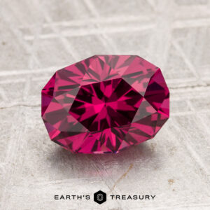 A 2.29-carat Tanga Garnet, featuring a faceted deep pink hue, is displayed on a textured light-gray surface with "EARTH'S TREASURY" written below.