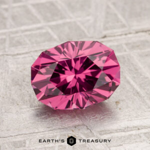 A 2.05-carat Tanga Garnet, oval-cut and faceted, rests on a textured gray surface with "Earth's Treasury" displayed at the bottom.