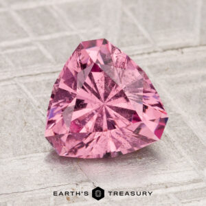 A 2.05-carat Tanga Garnet, faceted in a triangular cut and pink in color, is displayed on a textured surface with the text "Earth's Treasury" at the bottom.