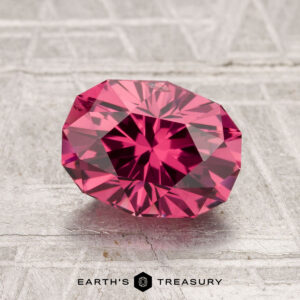 A 2.01-carat Tanga Garnet, faceted in an oval shape, rests on a textured light gray surface with "EARTH'S TREASURY" at the bottom of the image.