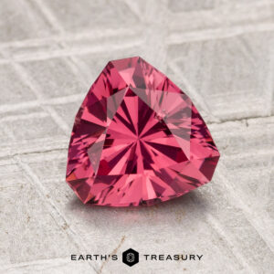 A 1.49-carat Tanga Garnet, cut in a triangular shape with a vivid pink hue, sits on a textured surface with the text "EARTH'S TREASURY" displayed below.