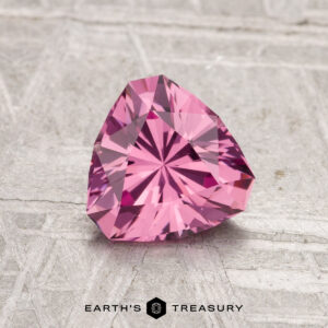 A 1.18-carat faceted triangular Tanga Garnet is displayed on a textured light gray surface with the text "EARTH'S TREASURY" below.