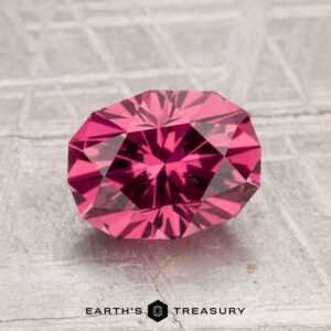 A 1.90-carat Tanga Garnet, faceted and deep pink, rests on a textured gray surface above the "EARTH'S TREASURY" text and hexagon logo.