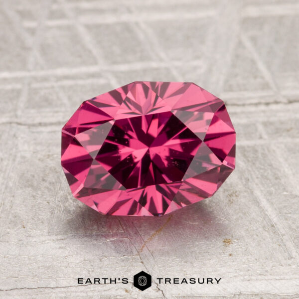 1.90-Carat Tanga Garnet A 1.90-carat Tanga Garnet, faceted and deep pink, rests on a textured gray surface above the "EARTH'S TREASURY" text and hexagon logo.