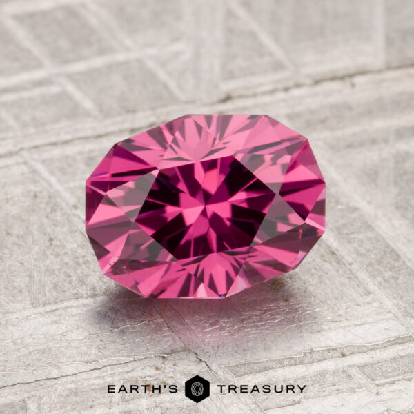 A 1.85-carat, oval-cut Tanga Garnet with a vibrant pink hue is displayed on a textured grey background, featuring the "Earth's Treasury" text and logo at the bottom.