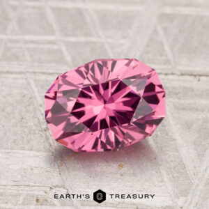 A 1.70-carat Tanga Garnet, cut in a faceted oval shape, rests on a textured gray surface with "Earth's Treasury" written below.