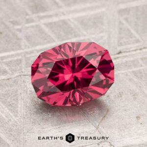 A 1.69-carat Tanga Garnet, faceted in an oval shape, is displayed on a textured metallic surface with the text "EARTH'S TREASURY" at the bottom.