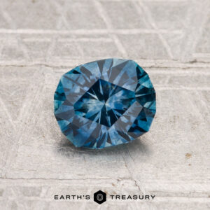 A 1.14-carat heated Montana sapphire is displayed on a textured grey surface, with the text "EARTH'S TREASURY" at the bottom.