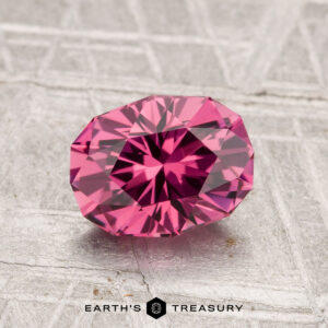A 1.55-carat Tanga garnet is displayed on a textured grey surface, with the "Earth's Treasury" logo beneath it.