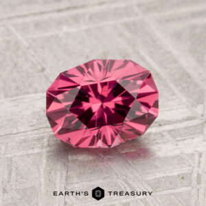 A faceted, round 1.48-carat Tanga garnet with a pink hue rests on a textured gray surface. The text "Earth's Treasury" appears at the bottom of the image.