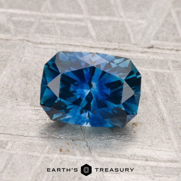 1.69-Carat Montana Sapphire (Heated) A 1.69-carat heated Montana sapphire with a rectangular cut is displayed on a textured gray surface, featuring the "Earth's Treasury" logo and text at the bottom.