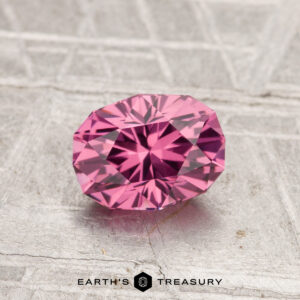 A 1.13-carat oval-cut Tanga Garnet with vibrant pink hues rests on a textured gray surface, featuring the Earth's Treasury logo at the bottom.