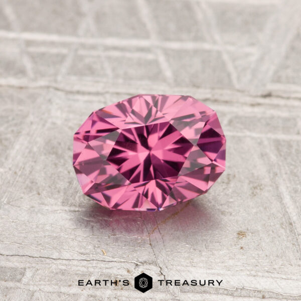 1.13-Carat Tanga Garnet A 1.13-carat oval-cut Tanga Garnet with vibrant pink hues rests on a textured gray surface, featuring the Earth's Treasury logo at the bottom.