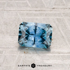 A radiant-cut, rectangular 1.09-carat heated Montana sapphire with a blue hue is displayed on a textured surface, with "EARTH'S TREASURY" shown beneath it.