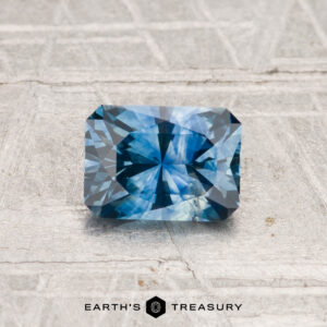 A rectangular, blue 0.90-carat heated Montana sapphire with intricate faceting rests on a textured surface. "EARTH'S TREASURY" appears at the bottom.