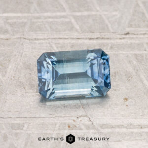 A rectangular, emerald-cut 1.08-carat heated Montana sapphire rests on a textured gray surface, with the Earth's Treasury logo below.