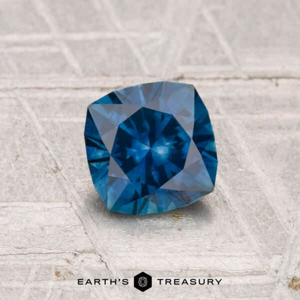 0.98-Carat Montana Sapphire (Heated) A 0.98-carat heated Montana sapphire, cushion-cut and blue, is displayed on a textured gray surface with "Earth's Treasury" at the bottom.