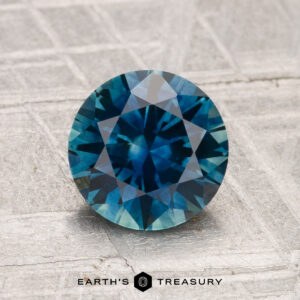 A 1.72-carat heated Montana sapphire, cut in a round brilliant shape, is displayed on a textured gray surface with "Earth's Treasury" at the bottom.
