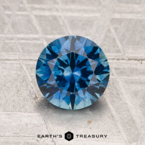 A 1.69-carat heated Montana sapphire, round brilliant-cut and blue, is displayed on a textured background with "EARTH'S TREASURY" at the bottom.