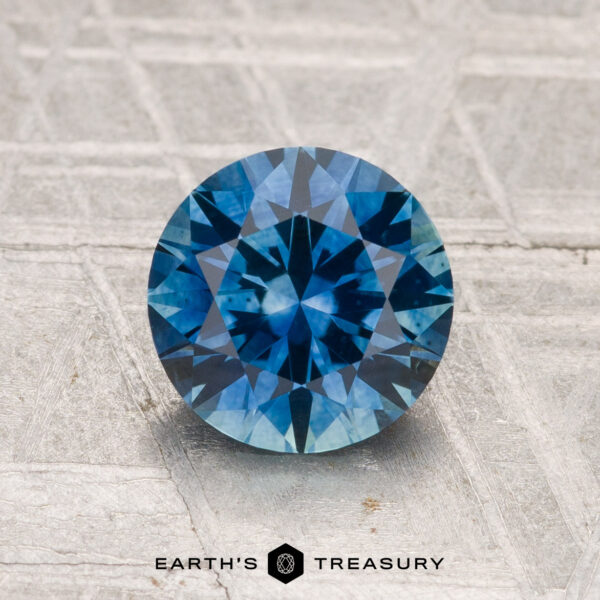1.69-Carat Montana Sapphire (Heated) A 1.69-carat heated Montana sapphire, round brilliant-cut and blue, is displayed on a textured background with "EARTH'S TREASURY" at the bottom.