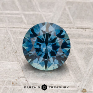 A 1.64-carat heated Montana Sapphire, round brilliant-cut and blue, displayed on a textured geometric surface with the "Earth's Treasury" logo at the bottom.