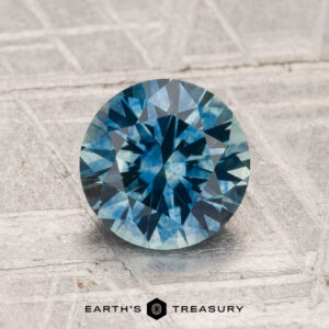 A round, faceted 1.63-carat Montana Sapphire (heated) with a blue-green hue rests on a textured silvery surface. The "Earth's Treasury" logo is visible at the bottom.