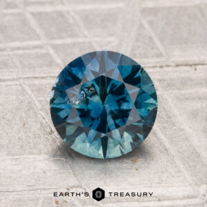 A 1.61-carat round brilliant-cut Montana Sapphire (heated) is displayed on a textured light gray surface with the “Earth’s Treasury” logo and text at the bottom of the image.