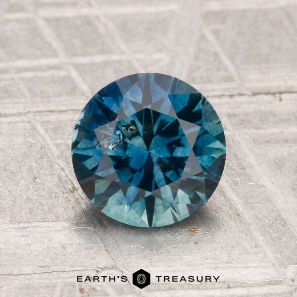 1.61-Carat Montana Sapphire (Heated) A 1.61-carat round brilliant-cut Montana Sapphire (heated) is displayed on a textured light gray surface with the “Earth’s Treasury” logo and text at the bottom of the image.