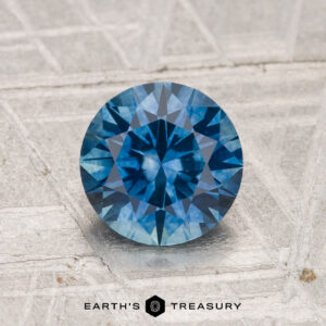 A 1.50-carat round brilliant-cut heated Montana sapphire is displayed on a textured metallic surface with the text "Earth's Treasury" at the bottom.
