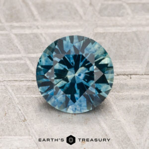 A 1.47-carat heated Montana sapphire, cut in a round blue-green facet, is displayed on a textured surface with "EARTH'S TREASURY" shown below.