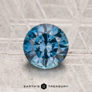 A 1.44-carat heated Montana Sapphire, round brilliant-cut, is displayed on a textured surface with "EARTH'S TREASURY" at the bottom.