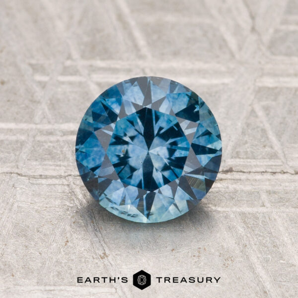 1.44-Carat Montana Sapphire (Heated) A 1.44-carat heated Montana Sapphire, round brilliant-cut, is displayed on a textured surface with "EARTH'S TREASURY" at the bottom.