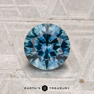A round, faceted 1.44-carat heated Montana sapphire in blue rests on a textured, light surface. "EARTH'S TREASURY" is displayed at the bottom of the image.