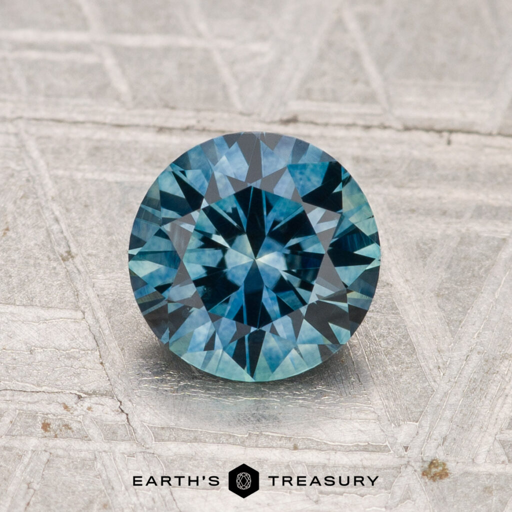 A 1.42-carat heated Montana sapphire, round brilliant-cut and blue, rests on a textured silver-gray surface with "EARTH'S TREASURY" at the bottom.