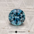 A 1.42-carat heated Montana sapphire, round brilliant-cut and blue, rests on a textured silver-gray surface with "EARTH'S TREASURY" at the bottom.