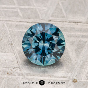 A 1.42-carat heated Montana sapphire, round brilliant-cut and blue, rests on a textured silver-gray surface with "EARTH'S TREASURY" at the bottom.