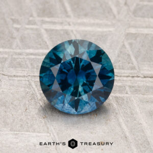 A round, faceted 1.38-carat Montana sapphire (heated) is displayed on a textured gray surface with the text "EARTH'S TREASURY" below it.