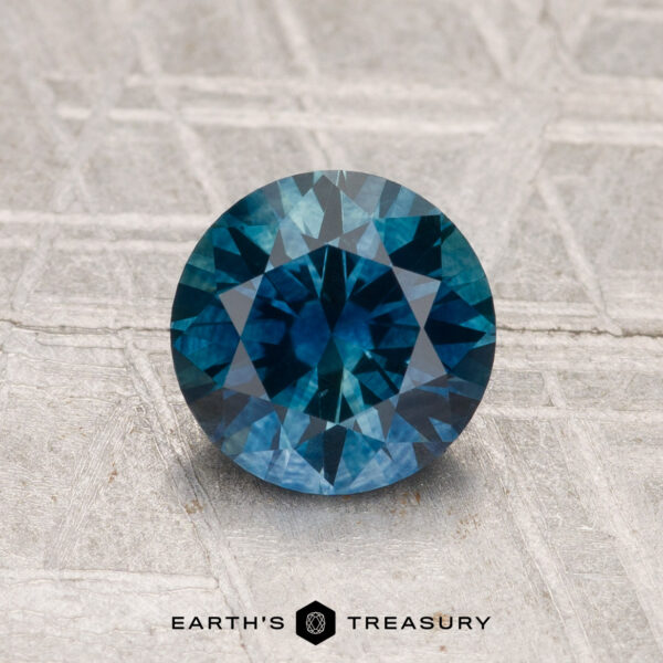 1.38-Carat Montana Sapphire (Heated) A round, faceted 1.38-carat Montana sapphire (heated) is displayed on a textured gray surface with the text "EARTH'S TREASURY" below it.