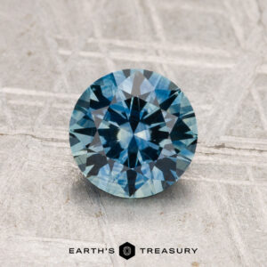 A round-cut 1.35-carat heated Montana sapphire with a faceted surface rests on a textured gray background. "Earth's Treasury" appears at the bottom.