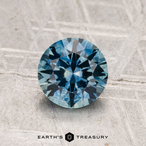 1.35-Carat Montana Sapphire (Heated) A round-cut 1.35-carat heated Montana sapphire with a faceted surface rests on a textured gray background. "Earth's Treasury" appears at the bottom.