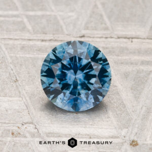 A 1.01-carat heated Montana sapphire, round brilliant-cut and blue in color, is showcased on a textured metallic surface with "EARTH'S TREASURY" displayed at the bottom.
