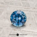 0.88-Carat Montana Sapphire (Heated), round brilliant-cut, blue gemstone displayed on a textured gray surface with "Earth’s Treasury" logo at the bottom.