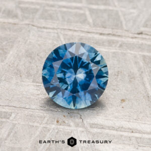 0.88-Carat Montana Sapphire (Heated), round brilliant-cut, blue gemstone displayed on a textured gray surface with "Earth’s Treasury" logo at the bottom.